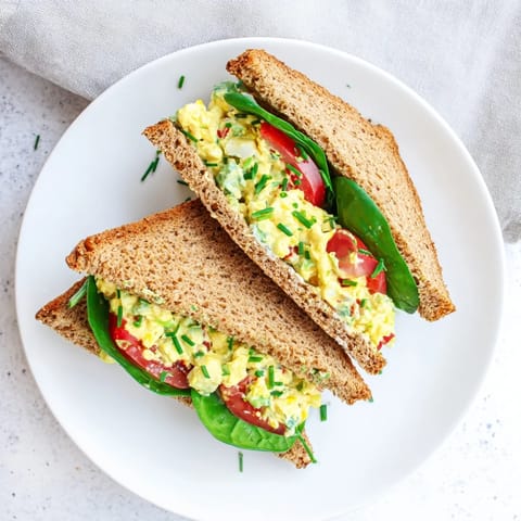 Ripe avocado egg salad on toasted whole grain bread, layered with fresh spinach and a slice of juicy red tomato.